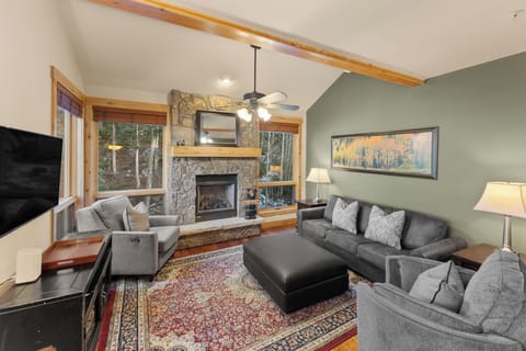 Cozy up by your stone fireplace while watching TV, surrounded by comfortable seating and forest views through large windows.