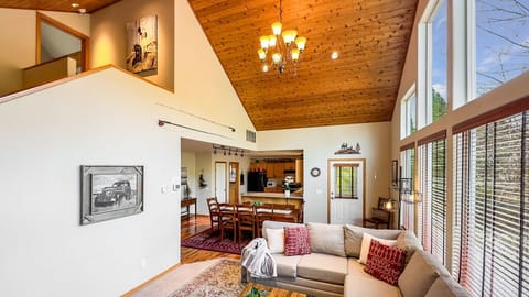 Vaulted chalet interior with dining for six, cathedral ceiling, and a wall of windows framing lush scenery.