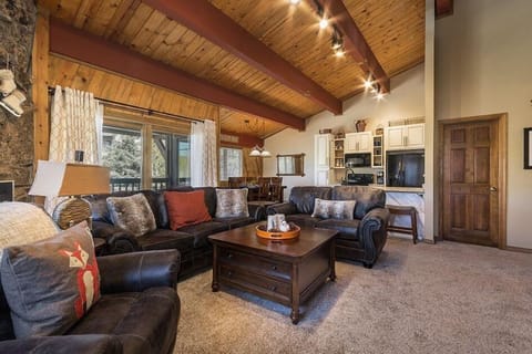 A spacious living room with wooden ceilings, dark sofas, a coffee table, and large windows. The background features a kitchen area with white cabinets and a dining table with chairs.