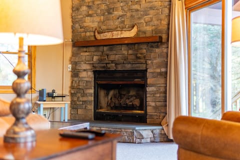 A gas fireplace is encased in a floor to ceiling stone facade with wood mantle and large flat screen TV.