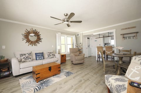 Relax in your bright, coastal-themed living space with comfortable seating, ceiling fan, and open layout connecting to your fully equipped kitchen.