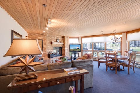 Unwind in your cozy living space with warm wood ceiling, crackling fireplace, and panoramic windows showcasing the city beyond.