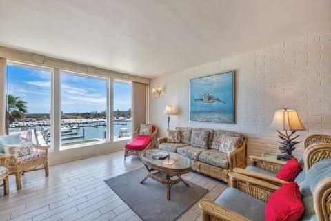 Relax in your bright living room with stunning marina views through floor-to-ceiling windows, featuring tropical wicker furniture and coastal artwork.