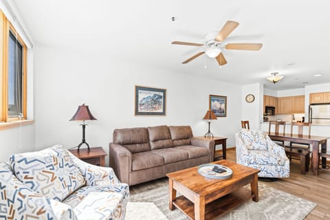 Relax in your open-concept living space with comfortable seating, mountain artwork, and a ceiling fan keeping you cool.