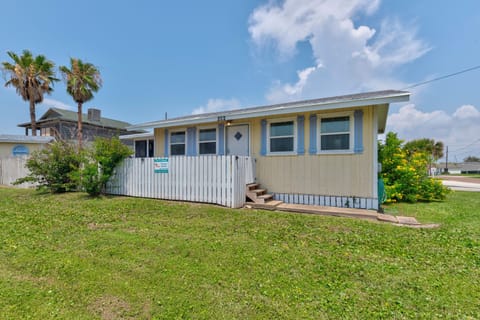 A charming coastal cottage nestled in a quiet neighborhood, featuring classic beachside architecture, a fenced yard, and tall palms swaying nearby under sunny skies.