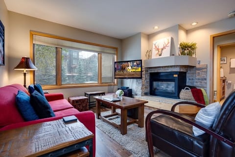 Cozy up by your stone fireplace with mountain views through large windows, surrounded by comfortable seating and warm lodge-style charm.