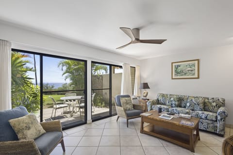 Relax in your bright, breezy living room with tropical-print seating, a ceiling fan, and stunning views through sliding doors to your private patio, gardens, and ocean beyond.