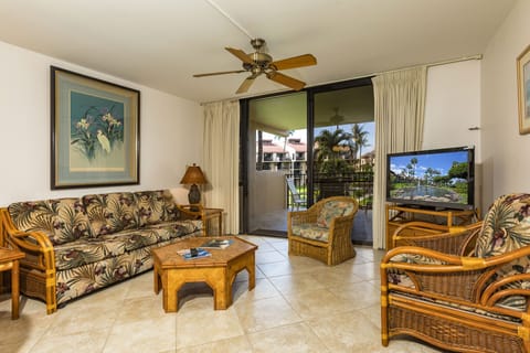 Your island getaway awaits in this bright, breezy living room with tropical furnishings, ceiling fan, and balcony doors opening to lush palms and serene poolside views.