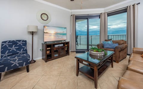 The living area features a large TV, comfortable seating, and access to a balcony with stunning views of the beach