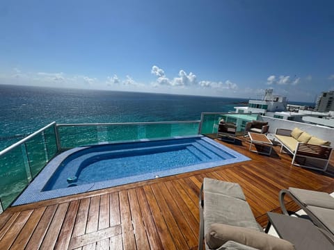 Rooftop deck with teak flooring, blue-tiled plunge pool, cushioned lounge chairs, and glass railing overlooking the ocean.