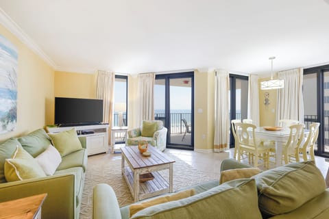Bright living space features green sofas, a TV, and dining space with a view of the gulf through large glass doors