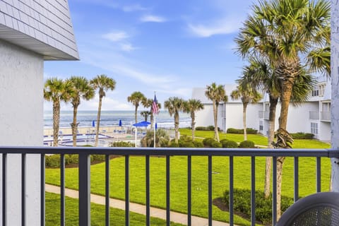 Balcony view overlooks manicured lawn, tall palm trees, a fenced pool area with blue umbrellas, and ocean beyond.