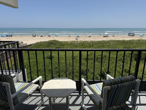 Second floor balcony with two striped cushioned chairs and a small white table facing green dunes and the ocean.