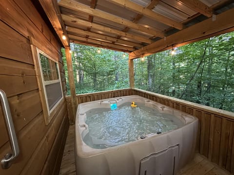 Covered porch holds bubbling hot tub with glowing blue light and floating yellow duck, surrounded by green trees.
