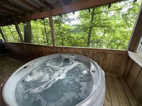 Bubbling round hot tub sits on a wood-paneled covered porch overlooking lush green trees.