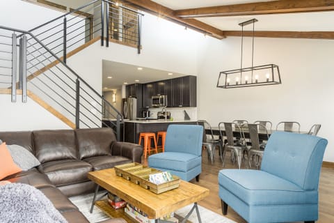 A modern living room with brown leather sofa, two blue chairs, a wooden coffee table, open kitchen, dining area with metal chairs, and staircase leading to an upper level.