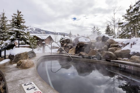 Outdoor hot spring pool surrounded by rocks and snow with steam rising, trees and mountain landscape in the background, and a sign indicating a depth of 3 feet 3 inches.