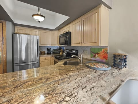 A kitchen with granite countertops, light wood cabinets, stainless steel refrigerator, black microwave, stovetop, and a colorful tile backsplash featuring flowers and a hummingbird.