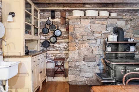 Stylish rustic kitchen with cream cabinetry, farmhouse sink, vintage décor, and a striking stone wall with a classic wood-fired stove—where charm meets functionality