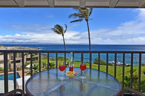 Oceanfront balcony featuring a four-seat dining table, lounge chairs, and sweeping views of turquoise waters, palm trees, and Kapalua Bay Villas.