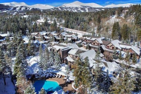 A stunning aerial view reveals a majestic winter retreat, where luxurious homes complement a vibrant blue pool, nestled among the mountains' snowy embrace.