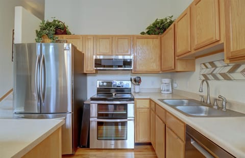 Modern kitchen with stainless steel appliances, wooden cabinets, and a dual sink. The countertop has a coffee maker and a knife set. Green plants adorn the top of the refrigerator and cabinets.