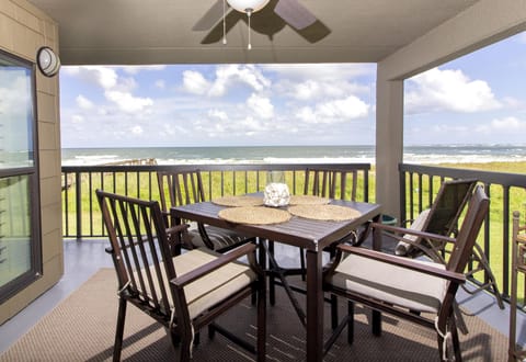 Covered balcony dining area with dark wood table, cushioned chairs, ceiling fan, and ocean views beyond railing.