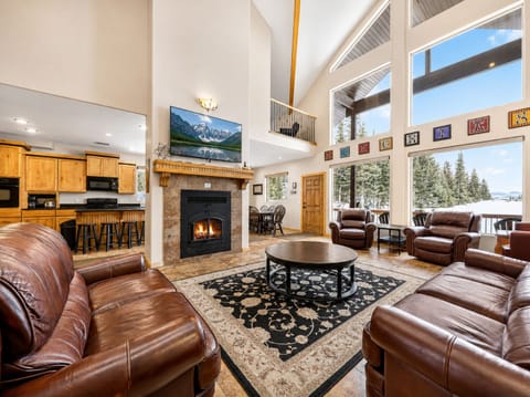 Unwind in your spacious living room with vaulted ceilings, cozy fireplace, plush leather seating, and stunning floor-to-ceiling mountain and forest views through expansive windows.