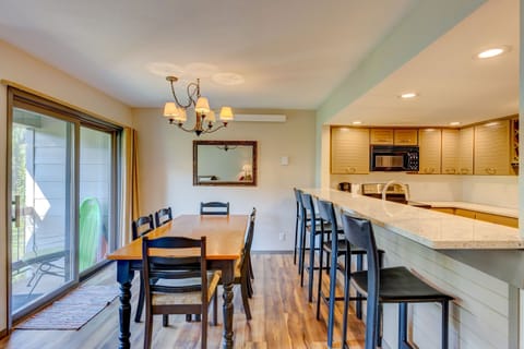 A dining area with a wooden table and six chairs, next to a kitchen with a breakfast bar and four bar stools. There's a chandelier above the table, a large mirror on the wall, and sliding glass doors on the left.