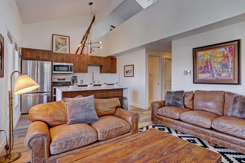A cozy living room with brown leather couches, a wooden coffee table, patterned rug, modern kitchen in the background, a staircase leading to an upper level, and colorful wall art.