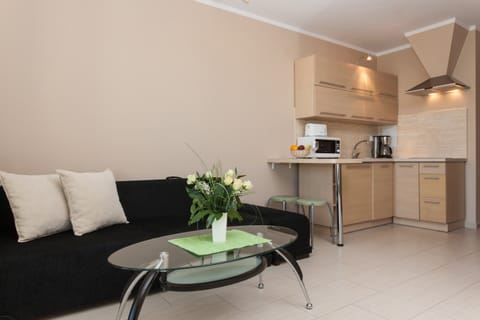 A small but modern living area with a black couch, two white pillows, and a round glass coffee table with a flower arrangement. In the background, you can see the kitchen area with light wood cabinets.