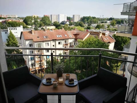 A small balcony with a table and two chairs, offering a scenic view of the city skyline, including rooftops and trees in the distance.