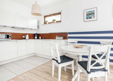 The kitchen is brightly coloured, with white cabinets and wooden accents. There is a dining table with four chairs against the wall with a window. A decorative blue stripe on the wall adds to the charm of the interior.