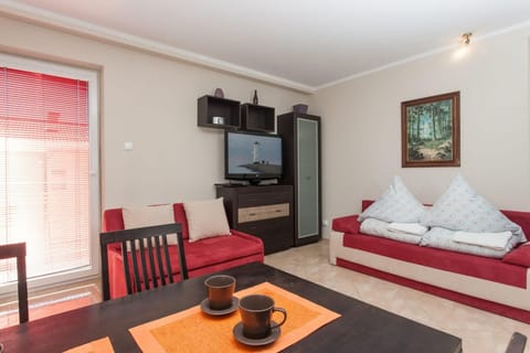 A cozy living space featuring two single beds with white bedding, positioned near a window with red blinds. A small table and dark wooden furniture add a warm, functional feel to the room.