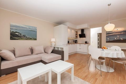 A spacious living room featuring a light-colored sofa set, a white coffee table, and a TV in the background. The room has a bright, airy feel with large windows and modern lighting.