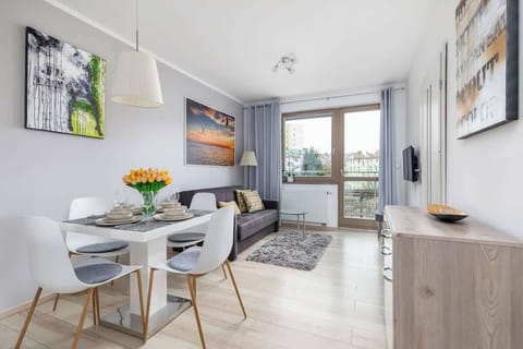 A dining area in a modern apartment with a round white table, four white chairs, and flowers as a centerpiece. Large windows provide natural light.