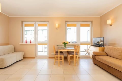 A bright living room with a beige sofa, large windows, and a small dining table. The space feels open and welcoming.