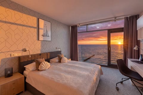 A cozy master bedroom is beautifully illuminated by a golden sunset visible through large floor-to-ceiling glass doors. The room features a double bed with neutral-toned linens and a sleek workspace desk nestled on the right.