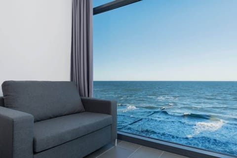 A modern living room with large glass windows, offering a stunning view of the sea. The room features a grey sofa and a light-colored coffee table, creating a serene, coastal ambiance.