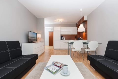 A bright, modern living room with two black sofas, a wooden coffee table, and light wood flooring. In the background, there is a dining table and chairs.