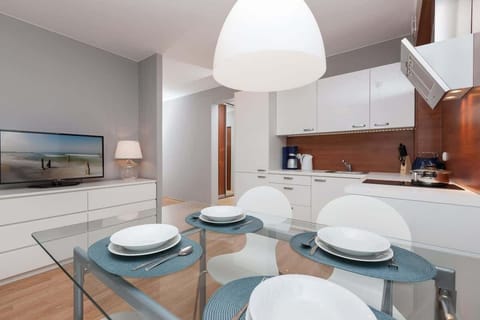 The kitchenette is bright and modern, with white cabinets and silver handles. In the foreground, there is a dining table with chairs, and a stylish lamp hangs above. The kitchen also features wooden accents near the countertop.