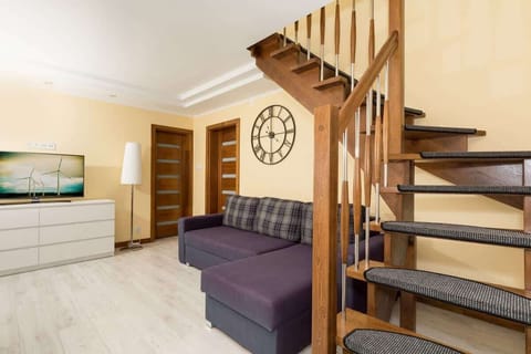 Bright, spacious living room with a chest of drawers on which stands the TV. To the right is a corner sofa bed above which is a village clock. In the right corner there are stairs leading upstairs.