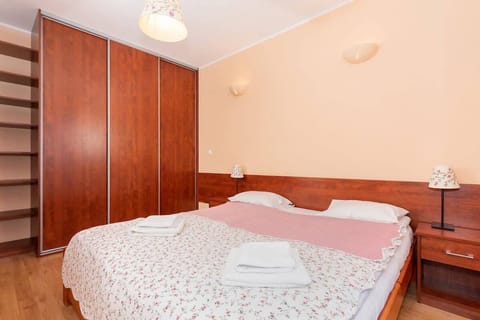 A bedroom with a double bed, dressed in a pink and white patterned bedspread. The room features a large wooden wardrobe and soft, peach-colored walls.