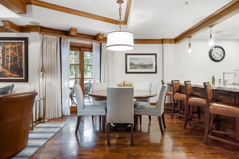 Dining area with round wood table, six gray upholstered chairs, and wood-trimmed ceiling beams.