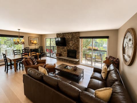 Living room with dark leather sectional, stone fireplace, wall-mounted TV, and sliding doors to balcony seating.