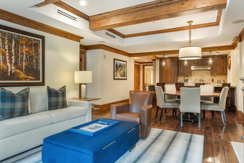 Living area with a cream sofa, plaid pillows, leather armchair, blue ottoman, and wood-beamed ceiling.