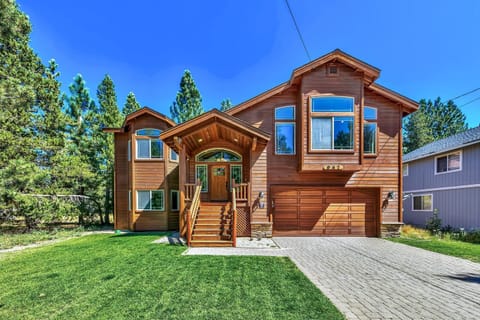 Wood-paneled exterior with front stairs, large windows, attached garage, and a green lawn bordered by tall pine trees.