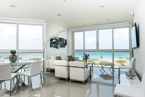 Bright modern living space with floor-to-ceiling windows framing pristine beach and turquoise ocean views, featuring sleek white furnishings and polished floors that reflect coastal sunlight.