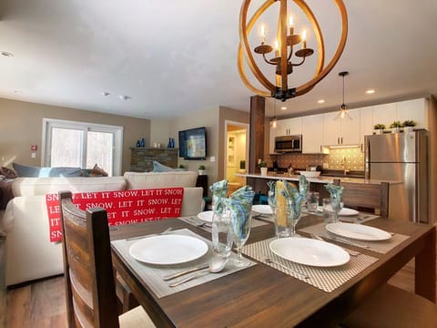 Modern farmhouse-style condo in Killington, Vermont. Main suite with its open kitchen featuring stainless appliances and an island, the dining area with seating for six, and the living room with a smart TV, sofa and gas fireplace seen at the back.