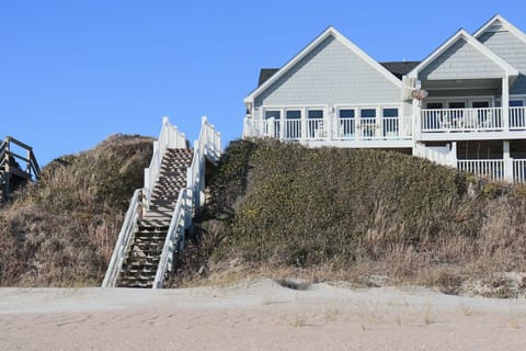 Charming beachfront home with private dune access stairs leads directly to pristine sandy shores beneath clear blue skies.